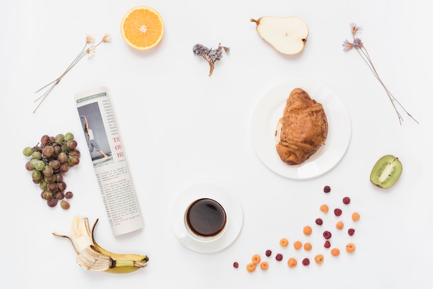 Foto gratuita enrollado periódico con taza de café; croissant y frutas sobre fondo blanco