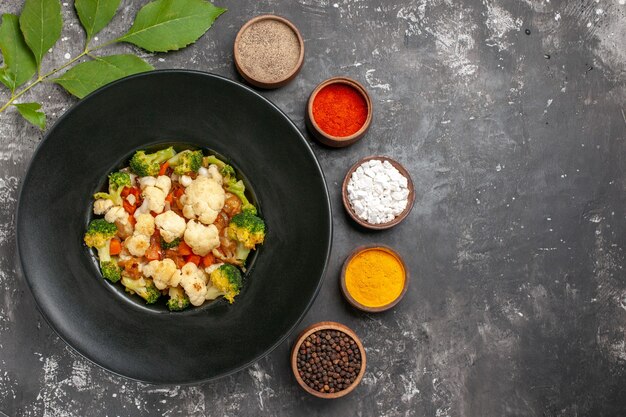 Ensalada de brócoli y coliflor de vista superior en placa ovalada negra diferentes especias en tazones pequeños en lugar libre de superficie oscura