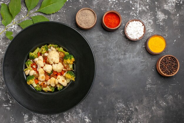 Ensalada de brócoli y coliflor de vista superior en plato ovalado negro diferentes especias en tazones pequeños pimientos de sal marina de cúrcuma en superficie oscura