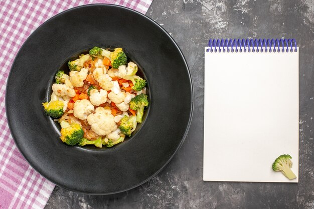 Ensalada de brócoli y coliflor de vista superior en plato ovalado negro mantel a cuadros rosa y blanco un cuaderno sobre superficie oscura