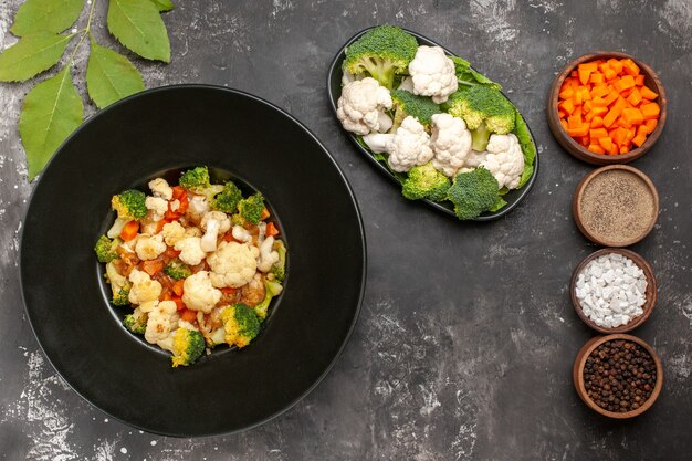 Ensalada de brócoli y coliflor de vista superior en tazón negro diferentes especias y zanahoria cortada en tazones brócoli crudo y coliflor en un plato sobre una superficie oscura