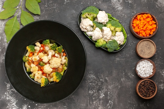 Ensalada de brócoli y coliflor de vista superior en tazón negro diferentes especias y zanahoria cortada en tazones brócoli crudo y coliflor en un plato sobre una superficie oscura