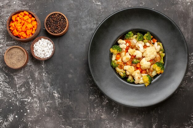 Ensalada de brócoli y coliflor de vista superior en tazón negro diferentes especias y zanahoria cortada en tazones sobre una superficie oscura con espacio de copia