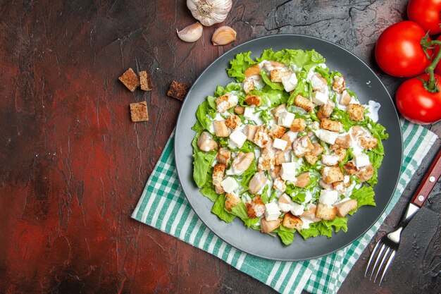 Ensalada César de vista superior en plato ovalado verde blanco servilleta a cuadros tomates tenedor sobre fondo rojo oscuro