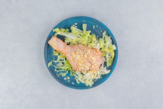 Ensalada y muslo de pollo en la placa de madera, sobre la superficie azul