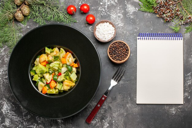 Ensalada de tomate verde de vista superior en placa ovalada un bloc de notas de tenedor especias sobre fondo oscuro