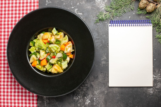 Ensalada de tomate verde de vista superior en plato ovalado mantel a cuadros blanco rojo un cuaderno sobre superficie oscura