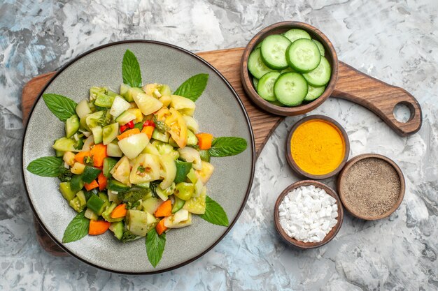 Ensalada de tomate verde de vista superior en tazón de fuente de placa ovalada con pepino cortado en tabla de cortar sobre fondo oscuro