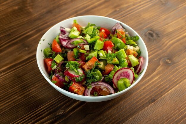 Ensalada de vegetales frescos, incluyendo pepinos en rodajas, tomates rojos, cebolla y otras cosas dentro de un plato blanco sobre la superficie rústica de madera