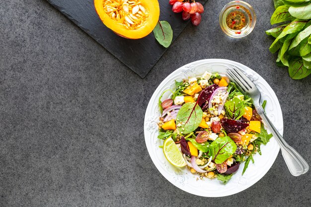 Ensalada de verduras frescas con remolacha, rúcula, cebolla morada, acedera, garbanzos, calabaza y uvas en un plato blanco sobre una mesa negra. Vista superior