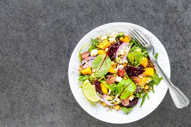 Ensalada de verduras frescas con remolacha, rúcula, cebolla morada, acedera, garbanzos, calabaza y uvas en un plato blanco sobre negro. Vista superior