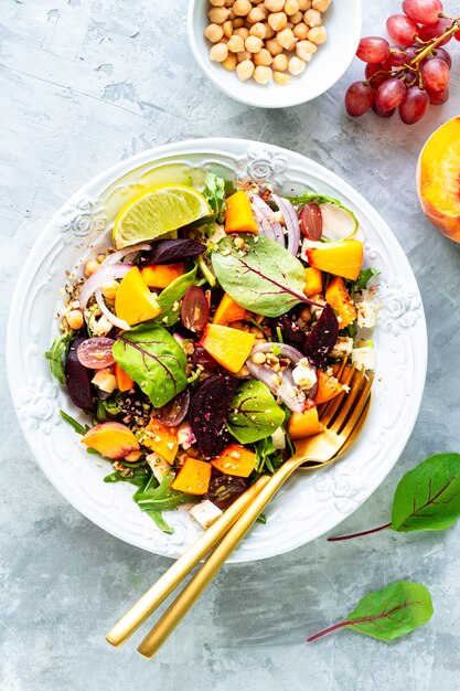 Ensalada de verduras frescas con remolacha, rúcula, cebollas rojas, acedera, garbanzos, melocotones y uvas en un plato blanco sobre piedra blanca. Vista superior