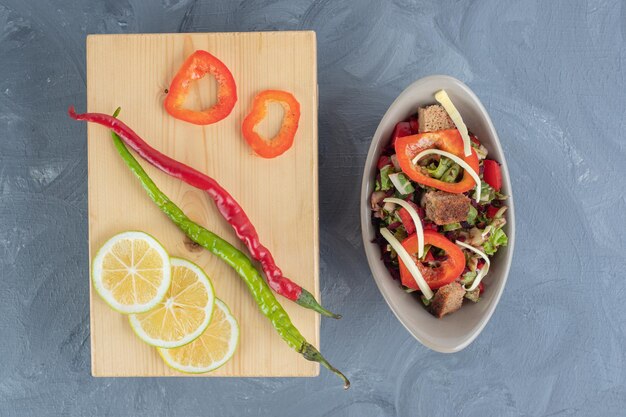 Ensalada de verduras junto a una tabla de madera con chiles y rodajas de limón y pimiento sobre mesa de mármol.