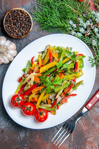 Ensalada de verduras vista superior en placa ovalada tenedor ajo pimienta negra sobre mesa rojo oscuro