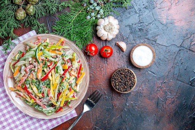 Ensalada de verduras de vista superior en placa sobre mantel tenedor sal y pimienta negra tomates al ajillo en mesa rojo oscuro