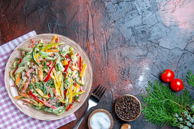 Ensalada de verduras vista superior en un plato sobre mantel tenedor sal y pimienta negra tomates al ajillo en el espacio libre de la mesa rojo oscuro