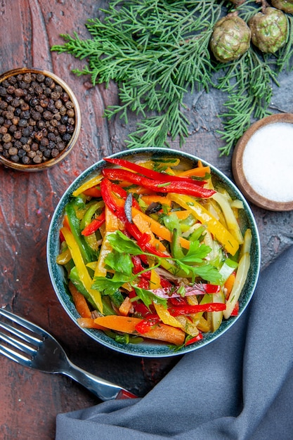 Ensalada de verduras de vista superior en un tazón de tenedor de pimienta negra de mantón azul ultramarino en mesa de color rojo oscuro