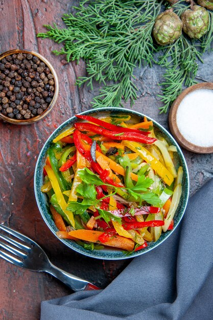 Ensalada de verduras de vista superior en un tazón de tenedor de pimienta negra de mantón azul ultramarino en mesa de color rojo oscuro