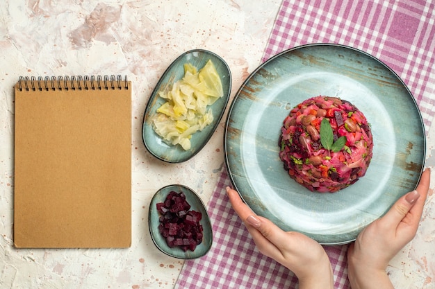 Ensalada de vinagreta de vista superior en plato ovalado en mano de mujer en tazones de mantel a cuadros blanco y morado con algunos blocs de notas en mesa gris claro