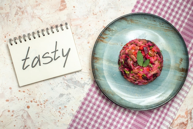 Ensalada de vinagreta de vista superior en plato ovalado mantel a cuadros blanco púrpura sabroso escrito en el cuaderno en la mesa gris claro