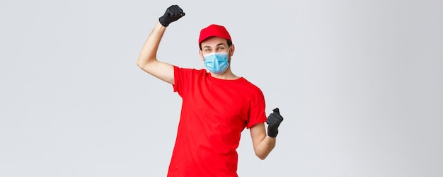 Entrega de abarrotes y paquetes, covid-19, concepto de cuarentena y compra. Mensajero alegre uniforme rojo, guantes y mascarilla, levantando las manos, cantando regocijo por las buenas noticias, lucha contra el coronavirus