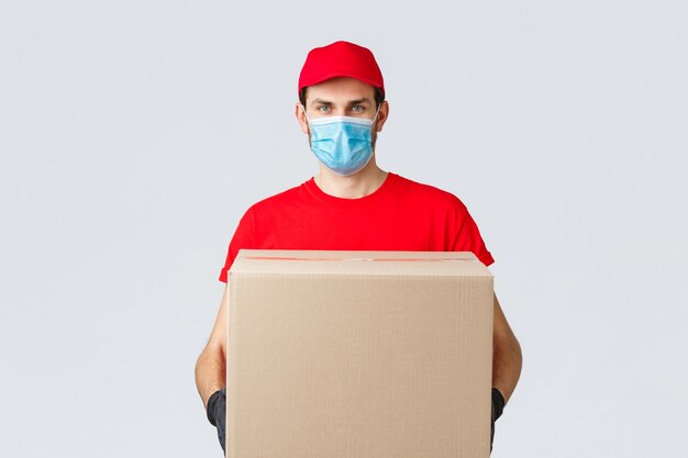 Entrega de abarrotes y paquetes, covid-19, concepto de cuarentena y compra. Mensajero serio con uniforme rojo, guantes y mascarilla protectora, entrega la caja del paquete a la casa del cliente durante el coronavirus