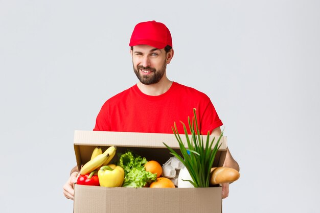 Entrega de abarrotes y paquetes, covid-19, concepto de cuarentena y compra. Mensajero sonriente guapo con uniforme rojo, guiño descarado como entrega de caja de comida, pedido en línea a la casa del cliente.