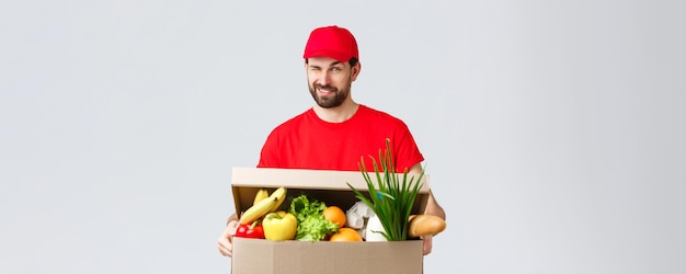 Entrega de comestibles y paquetes concepto de cuarentena y compras de covid19 Apuesto mensajero sonriente en uniforme rojo da un guiño descarado mientras entrega el pedido en línea de la caja de alimentos a la casa del cliente