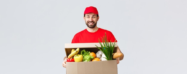 Entrega de comestibles y paquetes covid19 cuarentena y concepto de compras Repartidor sonriente en uniforme rojo trae el pedido del cliente sosteniendo el paquete de la caja de alimentos disfrutando de ayudar a las personas a transferir bienes