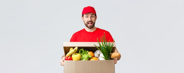 Entrega de comestibles y paquetes covid19 cuarentena y concepto de compras sonriente y guapo mensajero barbudo en uniforme rojo trae el paquete de alimentos pedido de comestibles al cliente en la caja se ve divertido