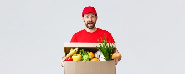 Entrega de comestibles y paquetes covid19 cuarentena y concepto de compras Sonriente y guapo mensajero barbudo en uniforme rojo trae el paquete de alimentos pedido de comestibles al cliente en la caja se ve divertido