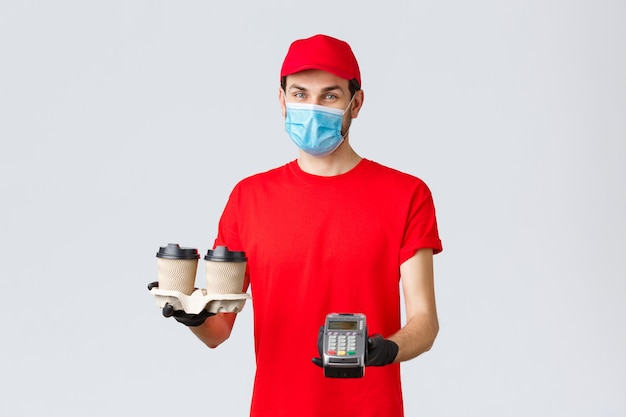 Entrega de comida para llevar, comida y abarrotes, concepto de pedidos sin contacto covid-19. Mensajero agradable en uniforme rojo, guantes y mascarilla, sosteniendo café para el cliente y terminal POS, fondo gris