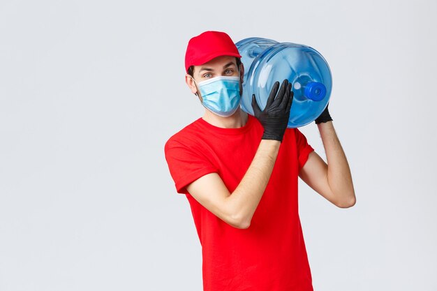 Entrega de comida para llevar, comida y abarrotes, concepto de pedidos sin contacto covid-19. Mensajero amistoso con uniforme rojo, gorra, mascarilla y guantes, con pedido de agua embotellada en el hombro