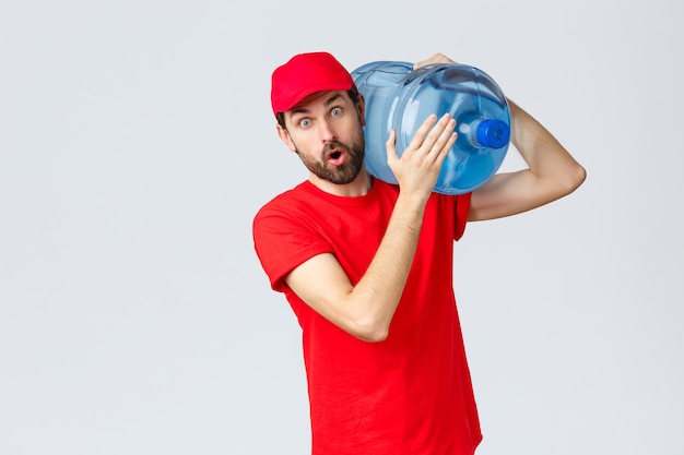 Entrega de comida para llevar, comida y abarrotes, concepto de pedidos sin contacto covid-19. Mensajero sorprendido con gorra de uniforme rojo y camiseta, boca abierta impresionado, sosteniendo agua embotellada en el hombro