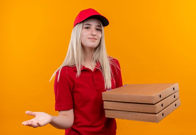 Entrega complacida joven vestida con camiseta roja y gorra con caja de pizza sosteniendo la mano a la cámara sobre fondo naranja aislado