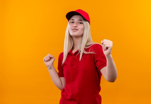 Entrega complacida joven vestida con camiseta roja y gorra mostrando gesto sí sobre fondo naranja aislado