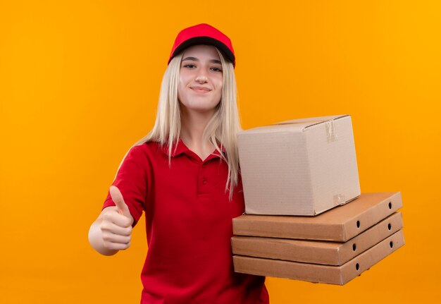 Entrega complacida joven vestida con camiseta roja y gorra sosteniendo cajas su pulgar hacia arriba sobre fondo naranja aislado