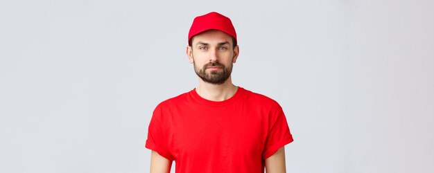 Entrega de compras en línea durante la cuarentena y el concepto de comida para llevar Joven y guapo mensajero barbudo en uniforme de servicio rojo con gorra y camiseta empleado que trabaja transfiriendo pedidos