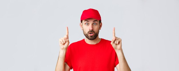 Entrega de compras en línea durante la cuarentena y el concepto de comida para llevar Sorprendido e impresionado mensajero repartidor con camiseta roja y gorra señalando con el dedo hacia arriba jadeando asombrado leer información interesante