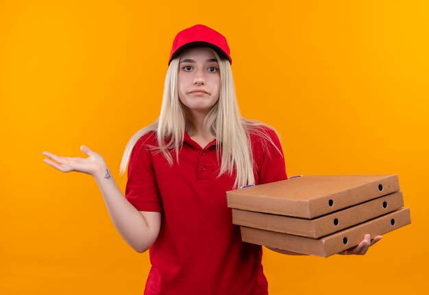 Entrega confusa joven vistiendo camiseta roja y gorra con caja de pizza en pared naranja aislada