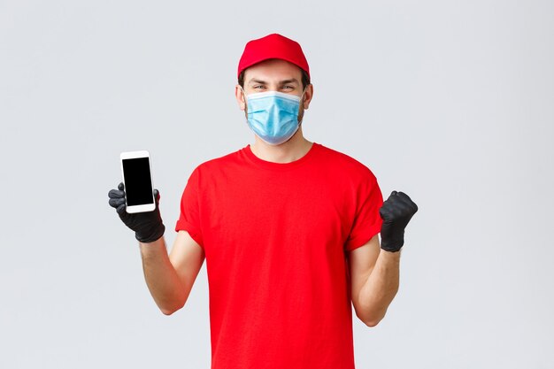 Entrega sin contacto, pago y compras en línea durante covid-19, autocuarentena. Regocijándose alegre mensajero con gorra de uniforme roja, camiseta celebrando una promoción increíble, mostrar la pantalla del teléfono inteligente, usar máscara