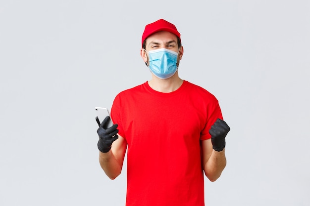 Entrega sin contacto, pago y compras en línea durante covid-19, autocuarentena. Regocijo, mensajero feliz en uniforme rojo, guantes y máscara, sonriendo, leer buenas noticias en la aplicación, teléfono móvil.