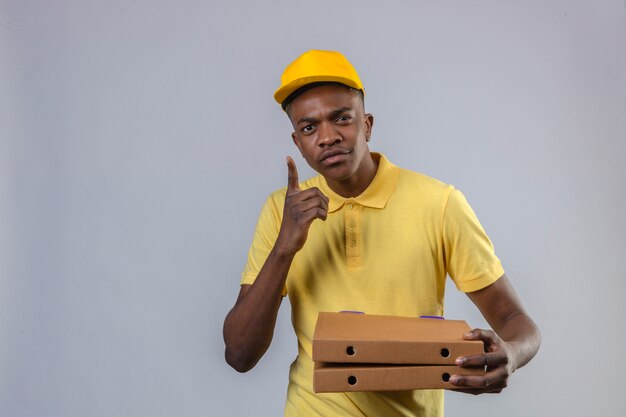 Entrega disgustado hombre afroamericano en camisa polo amarilla y gorra sosteniendo cajas de pizza apuntando con el dedo hacia arriba de pie