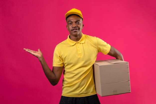 Entrega hombre afroamericano en camisa polo amarilla y gorra con paquete de caja presentando y señalando con la palma de la mano de pie en rosa aislado