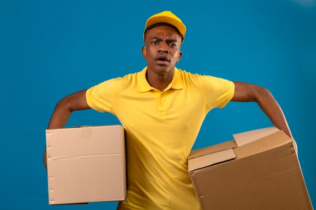 Entrega hombre afroamericano en camisa polo amarilla y gorra sosteniendo cajas de cartón con aspecto estresado conmocionado con cara de sorpresa enojado y frustrado de pie en azul aislado