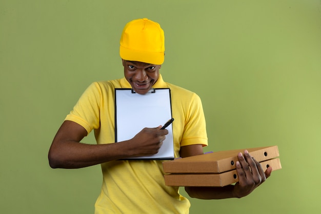 Entrega hombre afroamericano en camisa polo amarilla y gorra sosteniendo cajas de pizza y portapapeles pidiendo firma de pie en verde aislado