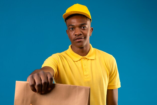 Entrega hombre afroamericano en camisa polo amarilla y gorra sosteniendo y extendiendo el paquete de papel sonriendo amable en azul aislado