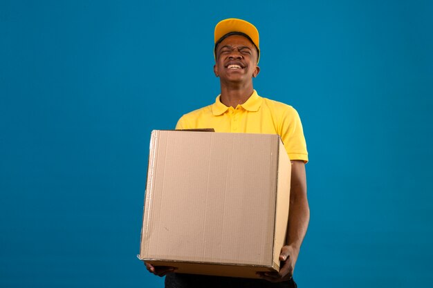 Entrega hombre afroamericano en camisa polo amarilla y gorra sosteniendo una gran caja de cartón pesado que se siente mal debido al gran peso en azul
