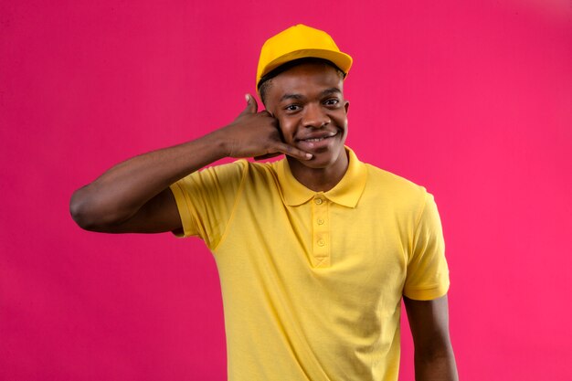Entrega hombre afroamericano en polo amarillo y gorra haciendo gesto de llamarme mirando confiado sonriendo alegremente en rosa