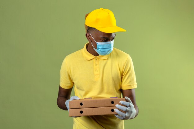 Entrega hombre afroamericano en polo amarillo y gorra con máscara protectora médica sosteniendo cajas de pizza mirando a un lado con cara seria de pie en verde